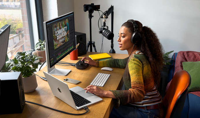 Una mujer con unos auriculares puestos trabaja desde su casa en un escritorio sobre el que se encuentra un MacBook Pro conectado a dos monitores externos y le habla a un micrófono