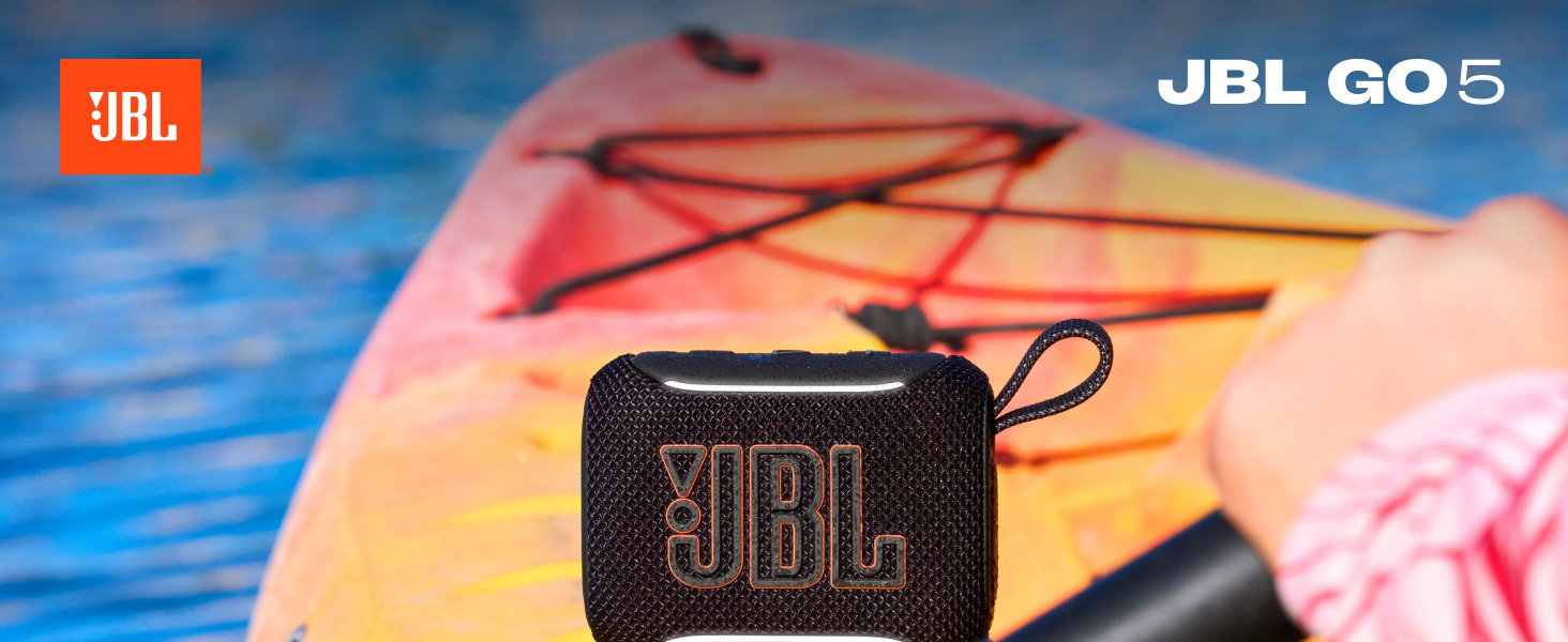 Il testo dice «JBL» e «JBL GO5". Altoparlante portatile nero su sfondo colorato sfumato con cavo, posizionato su una superficie a tema spiaggia.