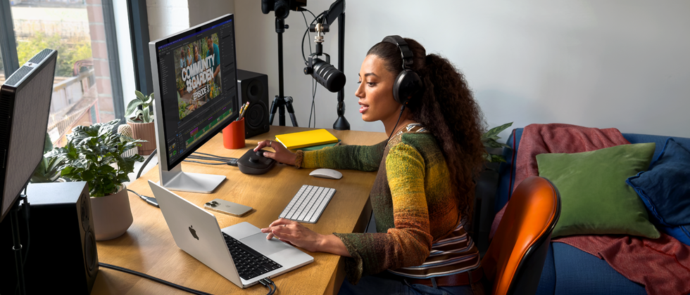 Una mujer con unos auriculares puestos trabaja desde su casa en un escritorio sobre el que se encuentra un MacBook Pro conectado a dos monitores externos y le habla a un micrófono