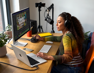 Una mujer con unos auriculares puestos trabaja desde su casa en un escritorio sobre el que se encuentra un MacBook Pro conectado a dos monitores externos y le habla a un micrófono